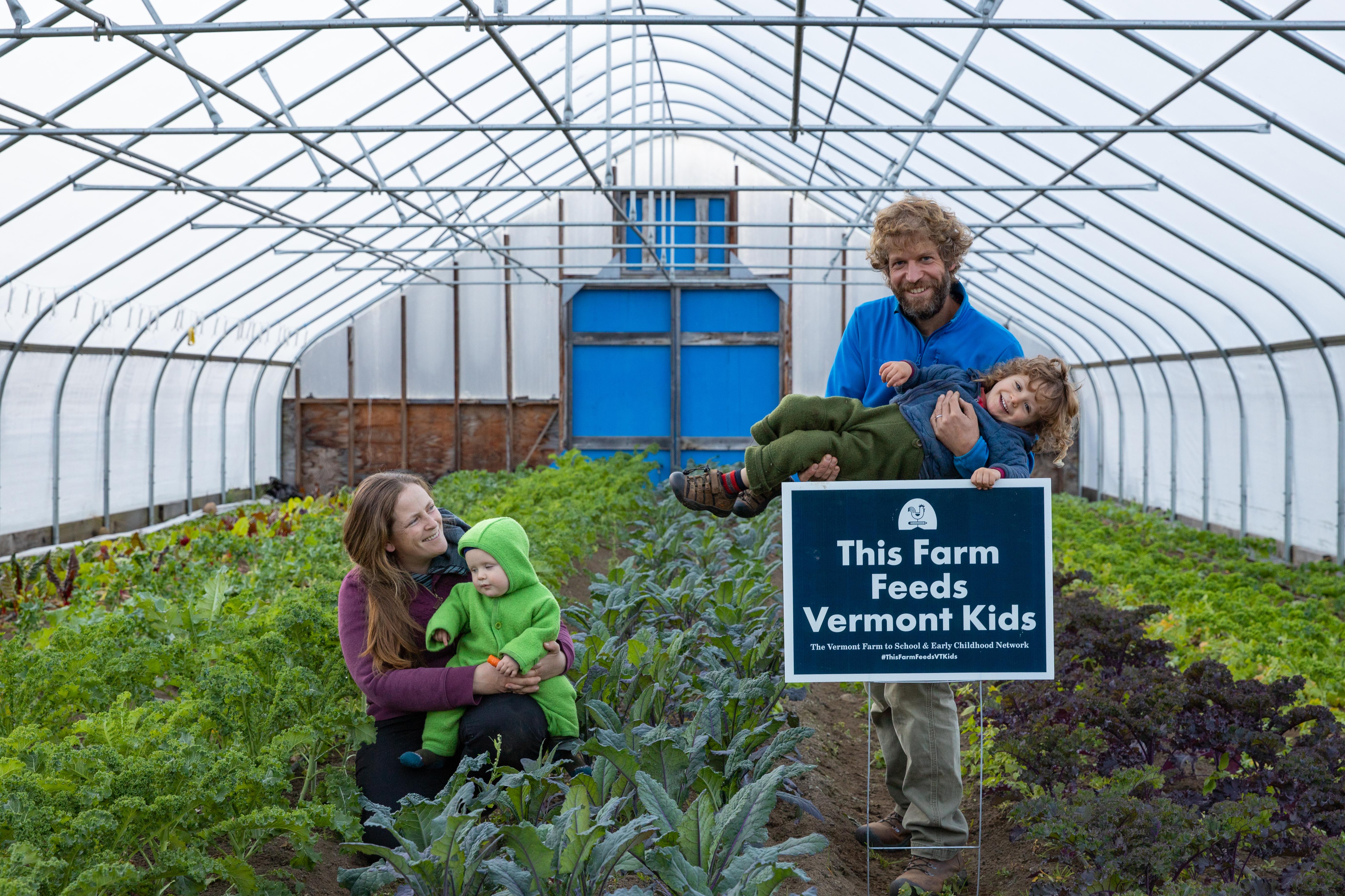 The farmers at Vermont's Long Winter Farm are pictured in their high tunnel filled with greens, holding their two children and standing with a sign that reads "this farm feeds Vermont kids"