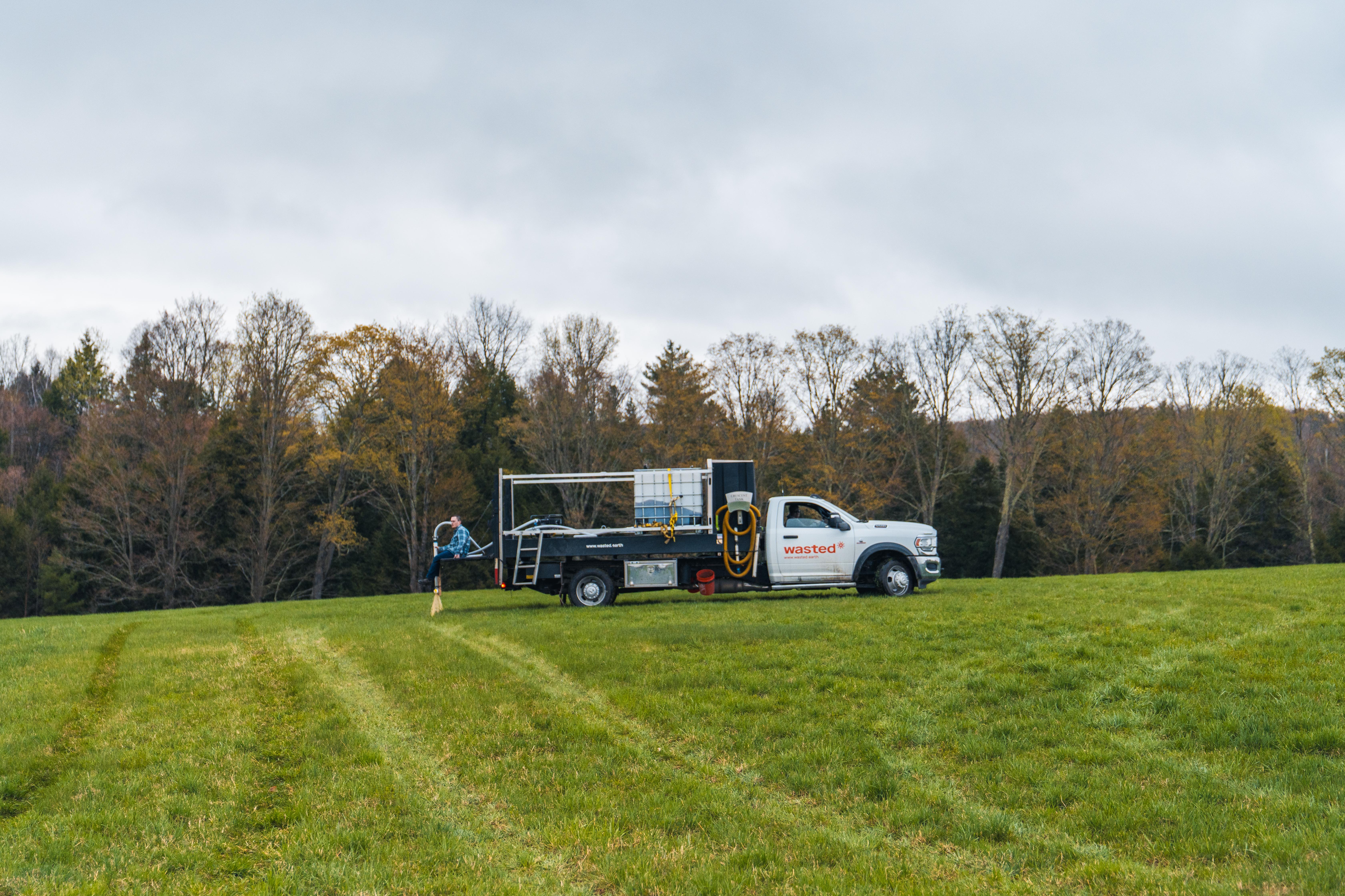 A Wasted* branded truck sits in a Vermont hay field, applying urine-derived fertilizer - open skies and liquid spraying out of a boom off the back of a truck.