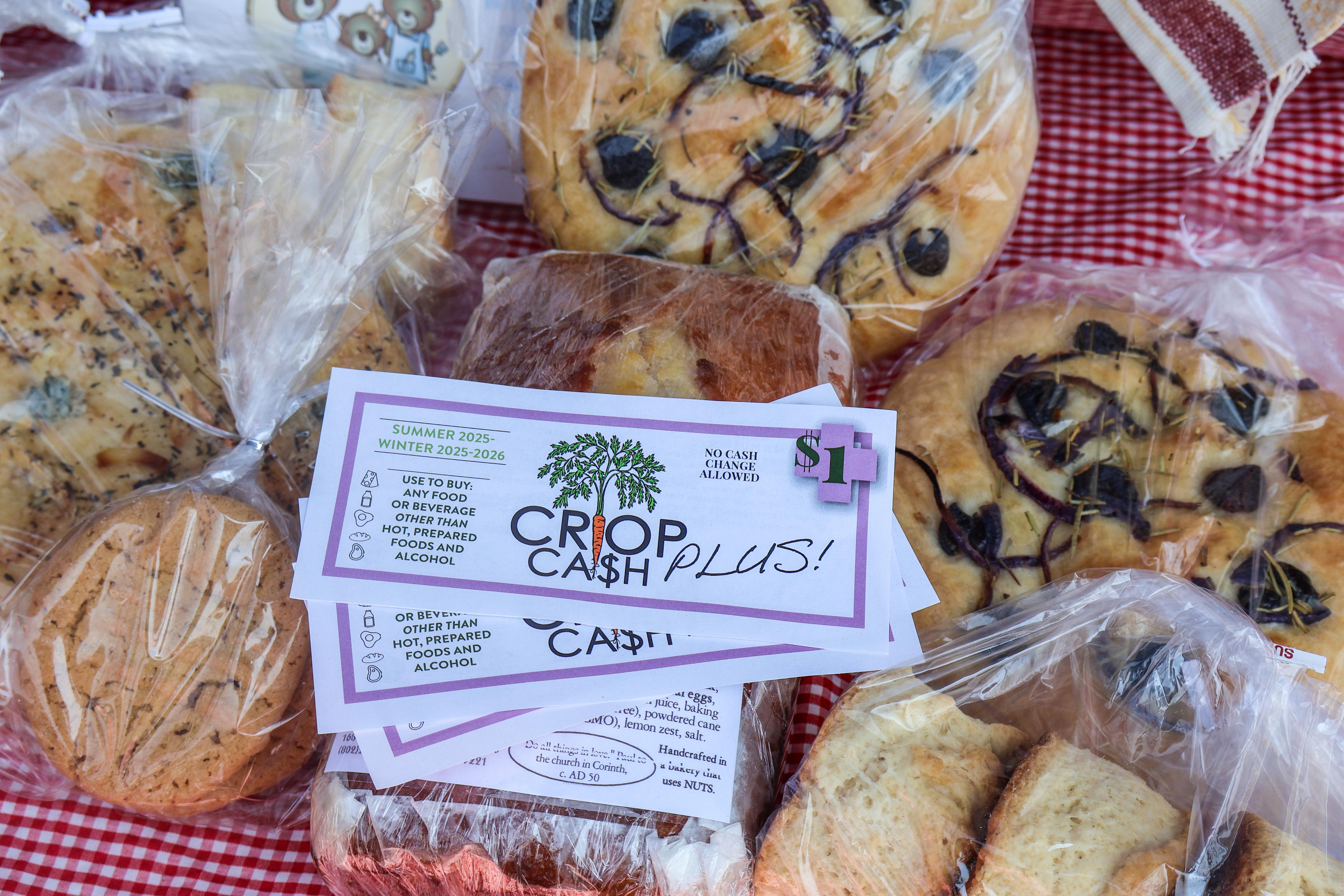 Crop Cash PLUS coupons fanned out in front of a farmers market booth of Crop Cash PLUS eligible items including bread and other baked goods from Three Bears Bakery