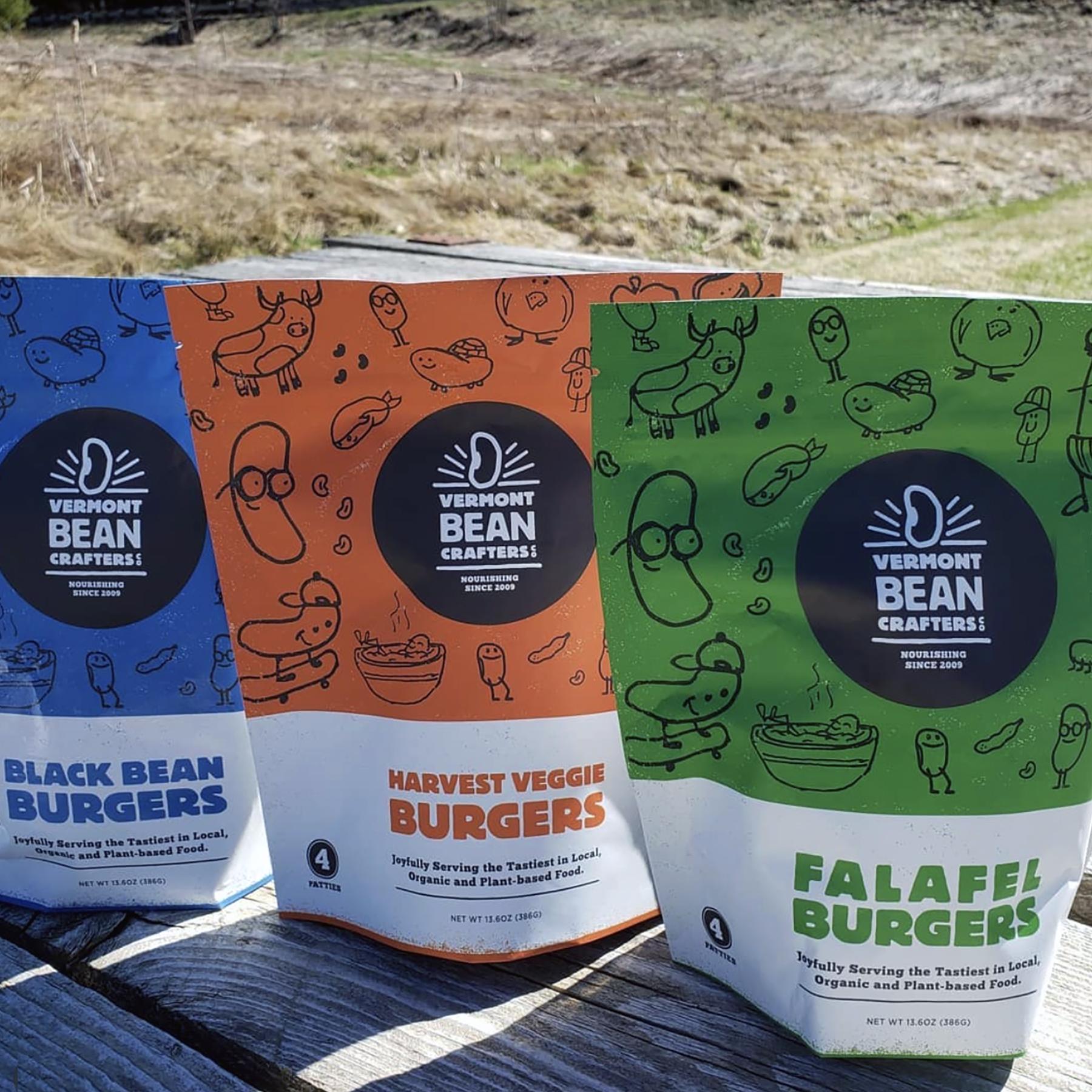 A lineup of three packages of Vermont Bean Crafters bean patties on a picnic table in an outdoor setting