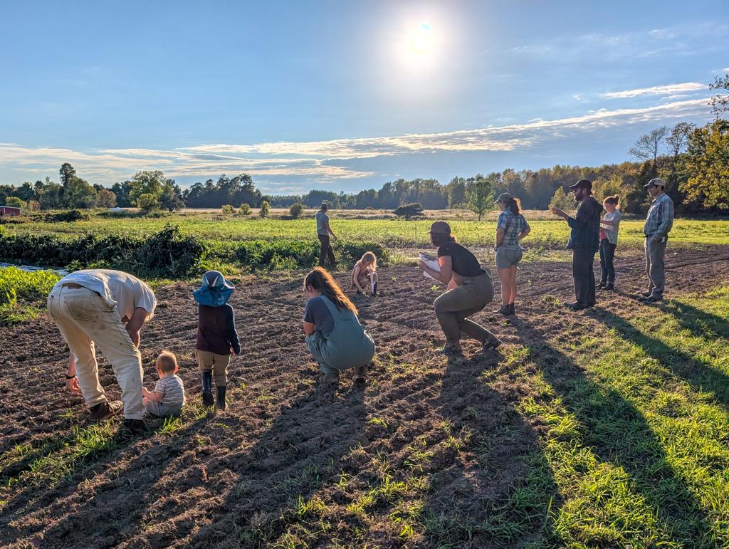 Eight adults and two kids stand dispersed on a small farm field in the sunshine.