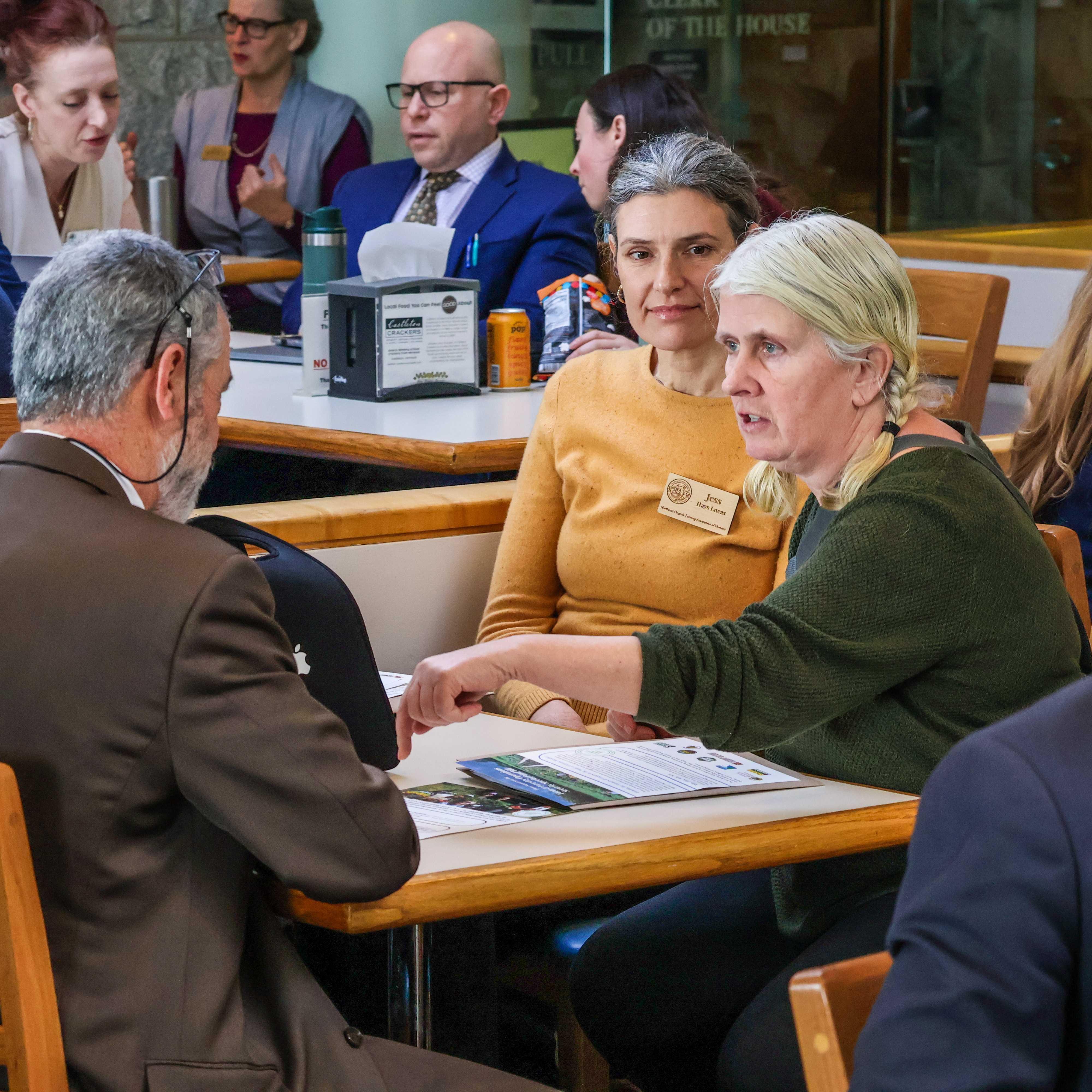 NOFA-VT farmer member Margaret Loftus of Crossmolina Farm and NOFA-VT Grassroots Organizer Jess Hays Lucas sit across the State House cafeteria table from Representative Mike Tagliavia, having a conversation about the The Farm and Forestry Operations Security Special Fund (S.60), while other legislators enjoy lunch in the background
