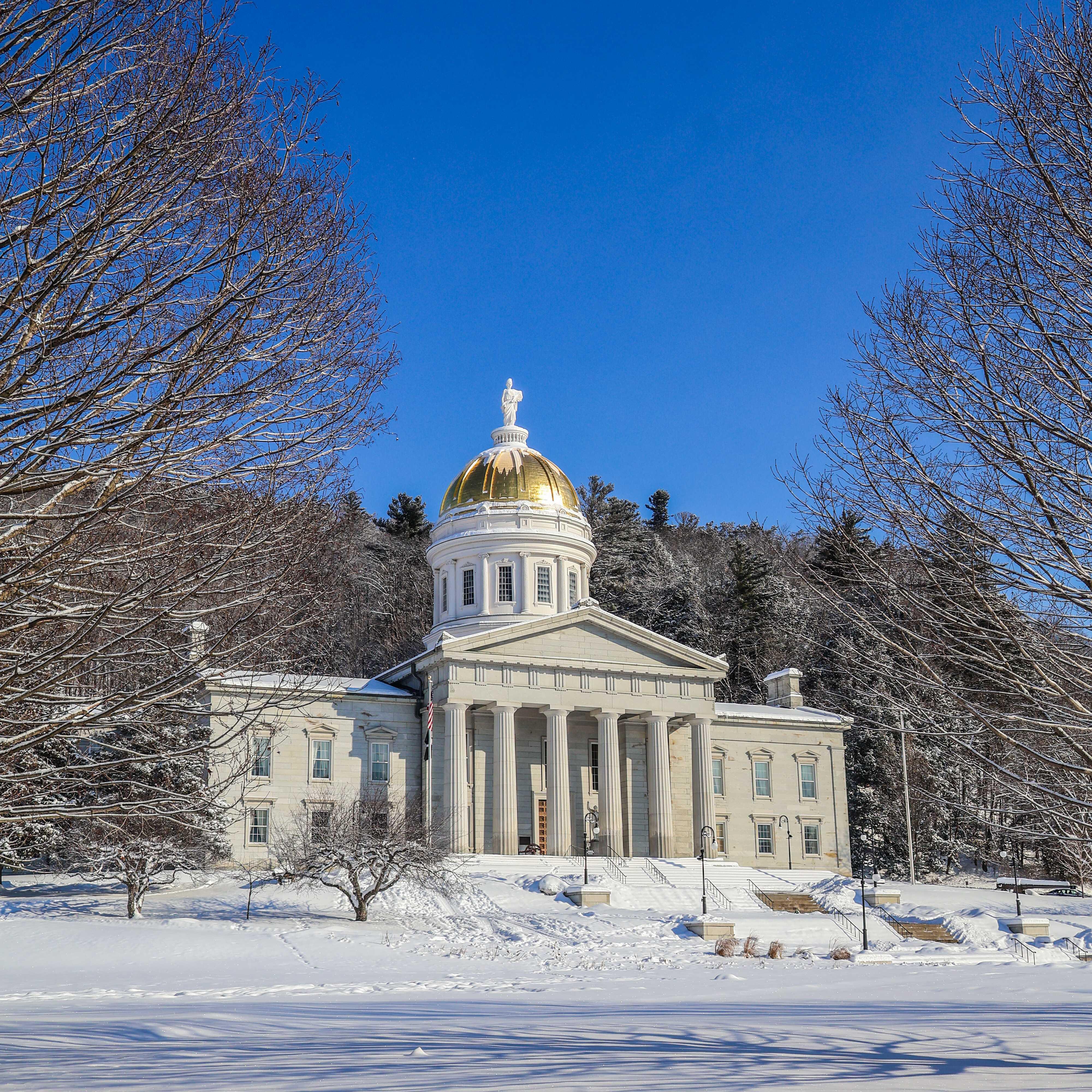 The State House in Montpelier on a bright winter day, with a vibrant blue sky and a blanket of white snow on the ground.
