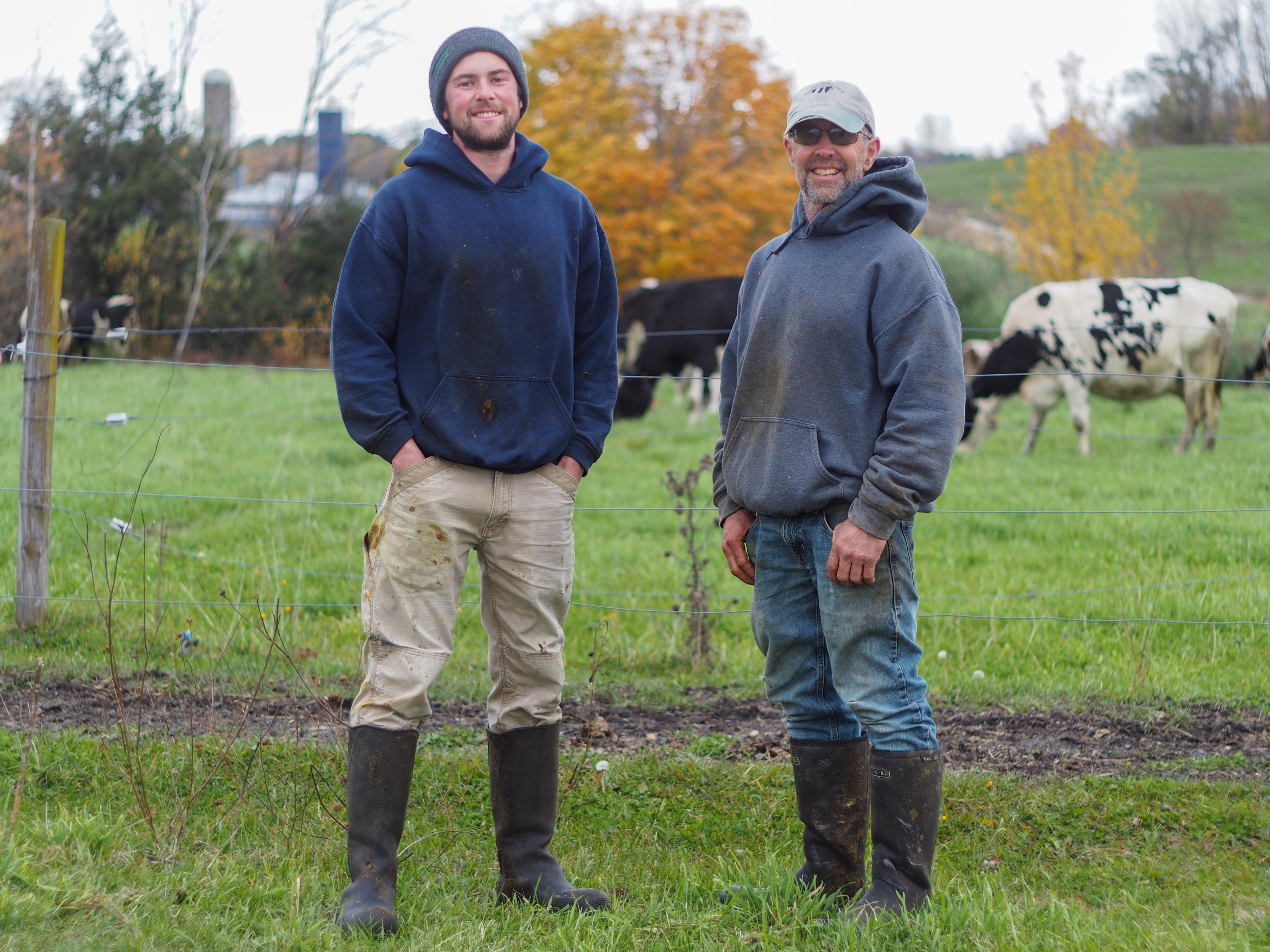 Matt and Guy Choiniere stand smiling in front of one of their cow pastures with cows grazing in the background