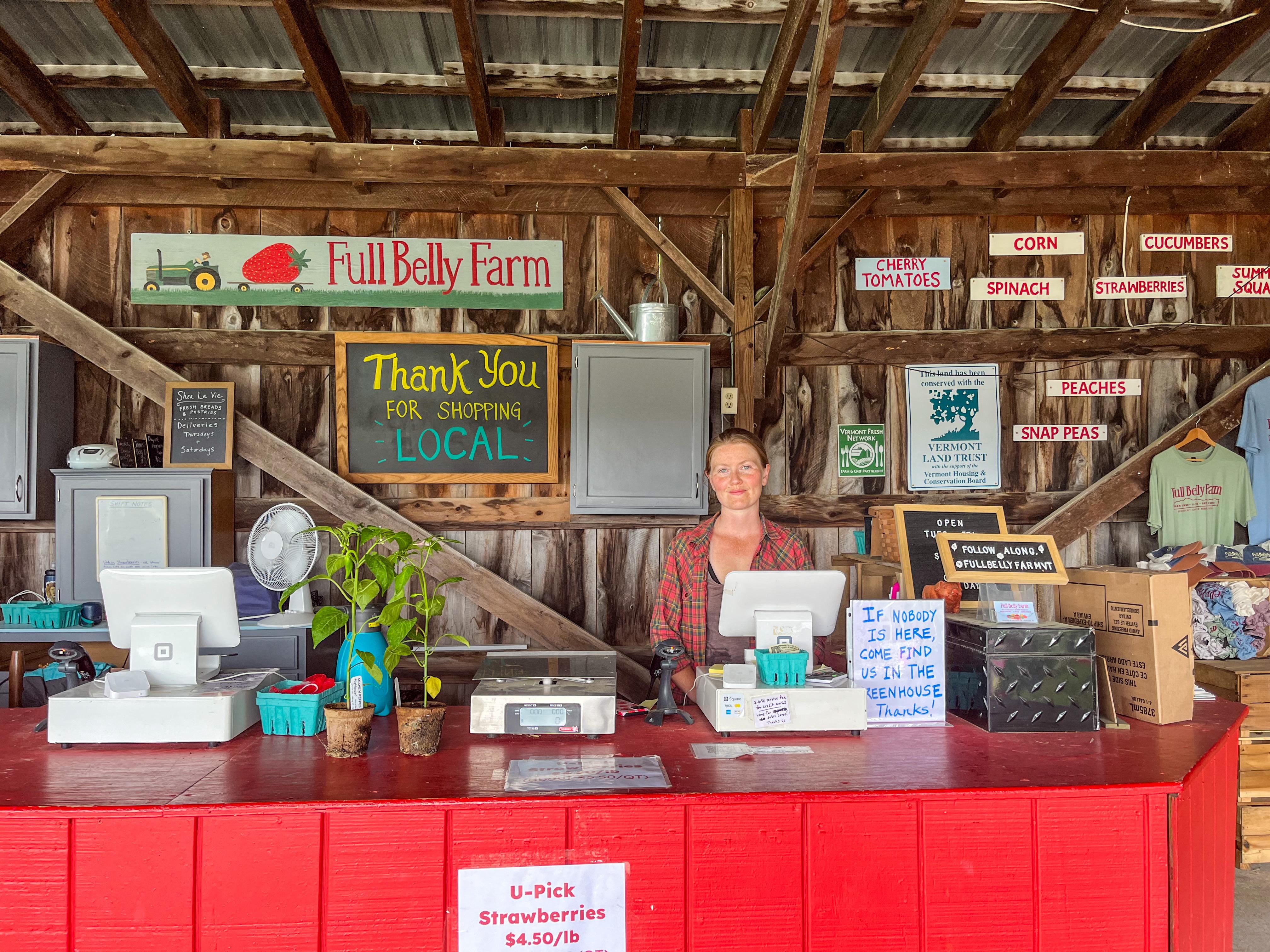 A smiling cashier is seen standing at the farm stand at Full Belly Farm, with an array of signs behind her welcoming visitors to their farm and thanking them for shopping local, a few plants on the counter, a POS system, and a few t-shirts hanging off to the right side.