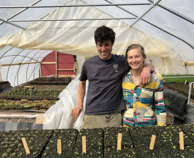 Gus and Mollie of Glinnis Hill Farm stand smiling in their high tunnel surrounded by trays of seedlings