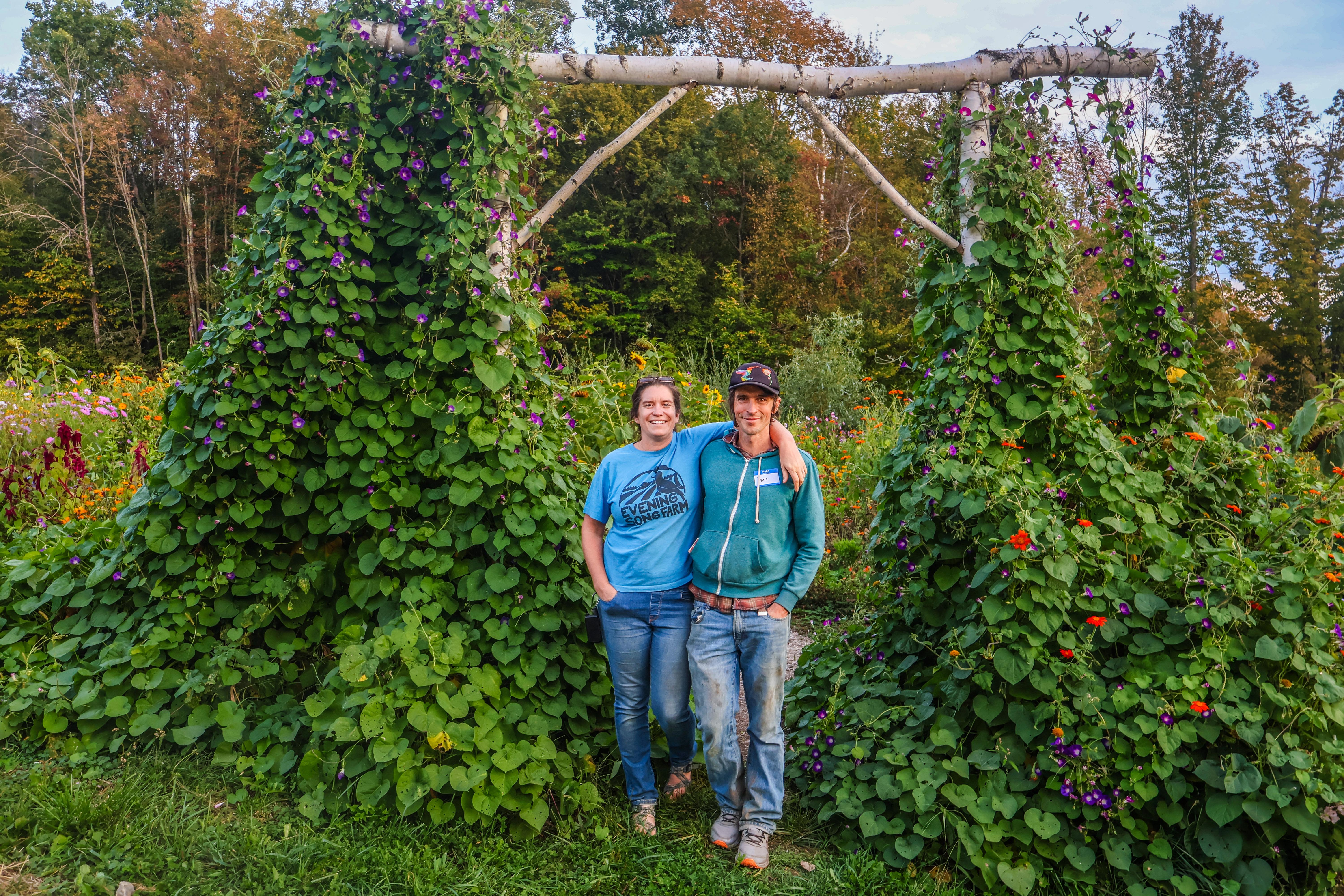 Farmers Kara and Ryan Fitzbeauchamp pose while smiling arm in arm under a wooden arbor covered in nasturtium vines at their farm