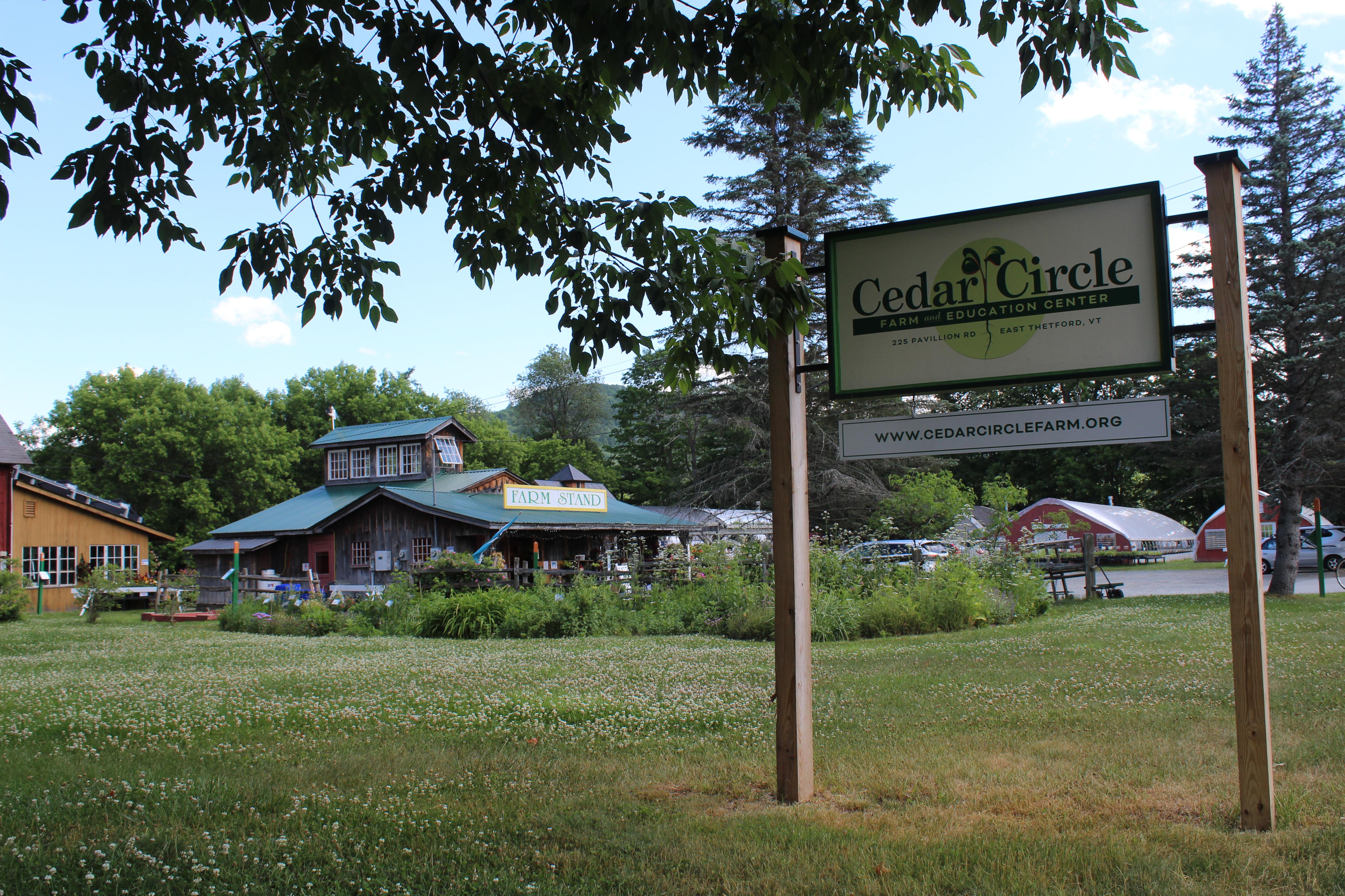 The roadside sign for Cedar Circle Farm in the foreground with the farm stand and several other outbuildings in the background