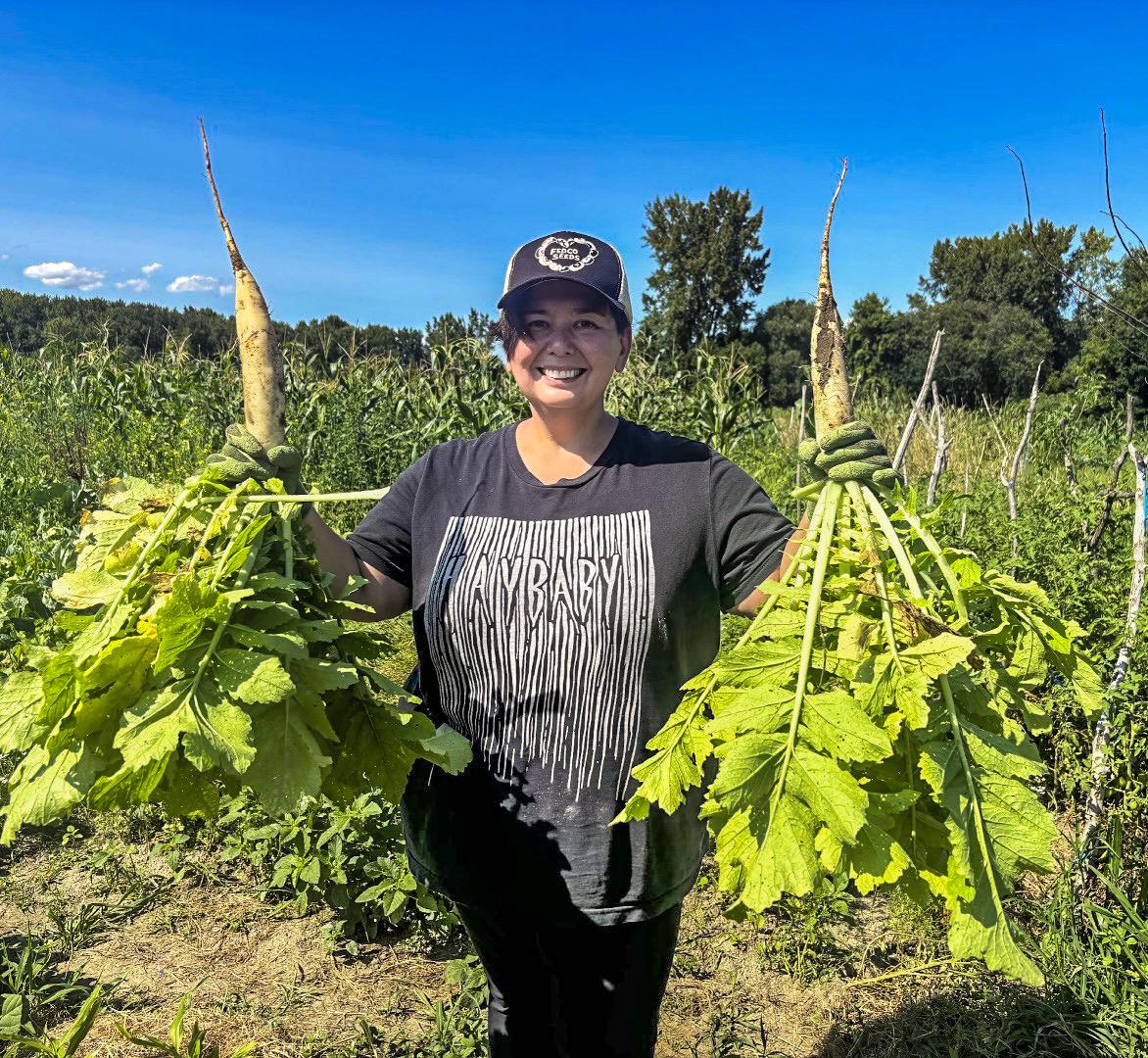 Alexis Yamashita stands smiling in a field while holding giant Daikon radishes in each hand