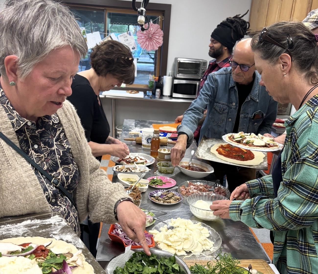 Five people top homemade pizzas around a kitchen island.