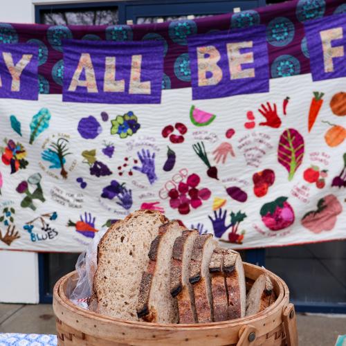 A basket of organic Red Hen bread sits on a table in the foreground with a colorful quilted banner reading "May All Be Fed" hanging in the background
