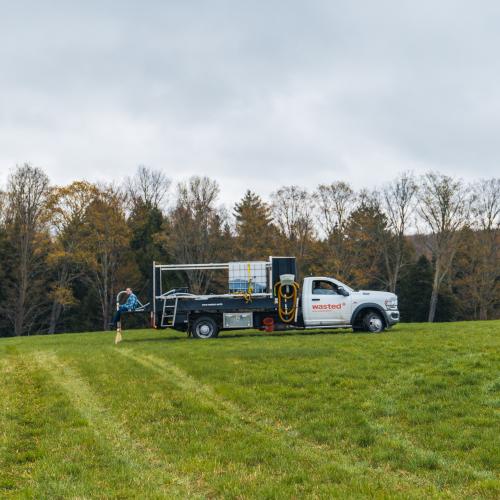 A Wasted* branded truck sits in a Vermont hay field, applying urine-derived fertilizer - open skies and liquid spraying out of a boom off the back of a truck.