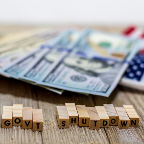 United States currency and an American Flag laying on a table in the background with small block letters reading "govt shutdown" in the foreground