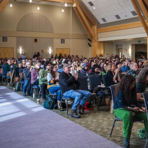 A crowd sits in a ballroom, looking up toward a speaker out of frame.