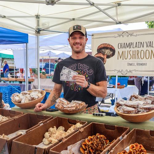 A representative from Champlain Vally Mushrooms stands at his farmers market booth holding Crop Cash coupons