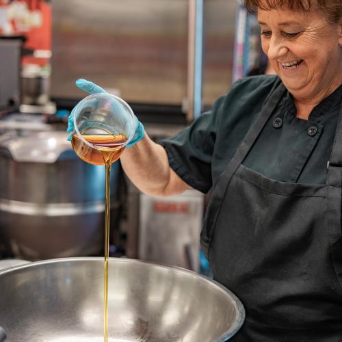 A Vermont school nutrition professional smiles while pouring Vermont maple syrup into a large stainless steel bowl in a school cafeteria