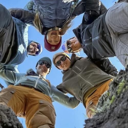 a photo taken from the ground, looking up at a huddle of 5 smiling faces linked arm-in-arm