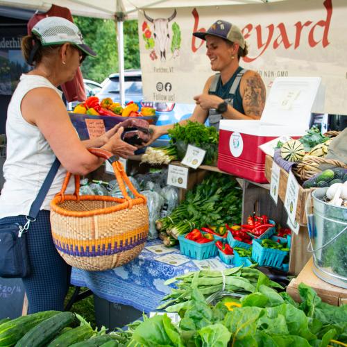 Hannah Doyle from Boneyard Farm is seen handling a transaction with a farmers market customer across a table abundantly filled with local, organic vegetables