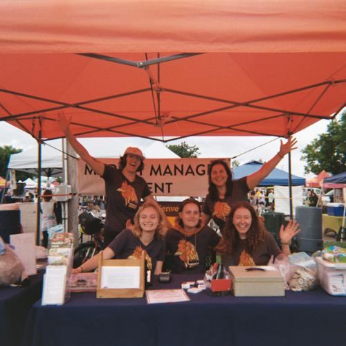 Staff pose behind the market manager table underneath an orange tent.