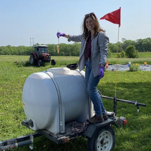 A person stands smiling while holding a test tube of urine over a plastic tank of urine fertilizer on a small trailer with fields and a tractor in the background