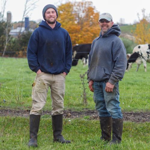 Matt and Guy Choiniere stand smiling in front of one of their cow pastures with cows grazing in the background