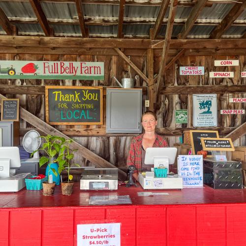 A smiling cashier is seen standing at the farm stand at Full Belly Farm, with an array of signs behind her welcoming visitors to their farm and thanking them for shopping local, a few plants on the counter, a POS system, and a few t-shirts hanging off to the right side.