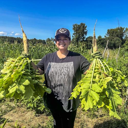 Alexis Yamashita stands smiling in a field while holding giant Daikon radishes in each hand