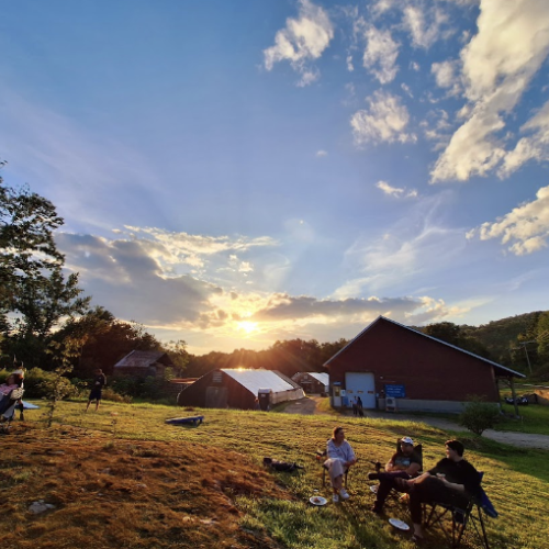 A beautiful sunset scene at Maple Wind Farm's Fried Chicken & Pickin' community event, with their farmstand and a vibrant sunset in the background and people enjoying the festivities in the foreground