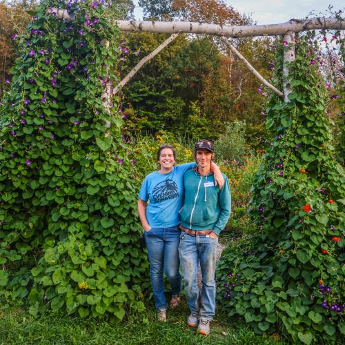 Kara and Ryan Fitzbeauchamp of Evening Song Farm stand smiling, arm in arm, underneath a wooden arbor covered in nasturtium vines.