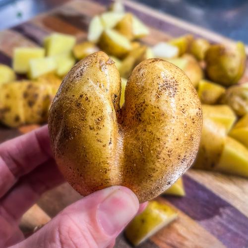 A heart-shaped potato is held close to the camera lens above a cutting board filled with diced potatoes.