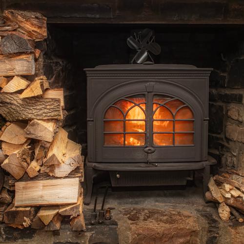 A big stack of wood next to a woodstove with a glowing orange fire visible through the glass paned door